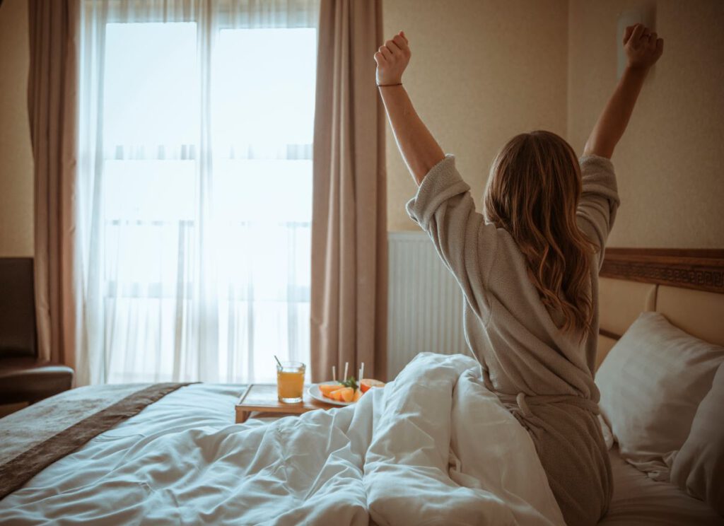 łózko w hotelu kobieta sie przeciąga na łóżku ma sniadanie na tacy sok pomarańczowy 